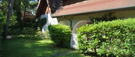 A house with a sloped roof, a front porch, and a well-maintained garden with a white fence and green bushes.