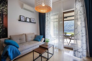 Ein Wohnzimmer mit einer Couch, einem Couchtisch, einer Hängeleuchte, einem an der Wand befestigten Regal mit gerahmten Bildern und Blick durch ein Fenster.
