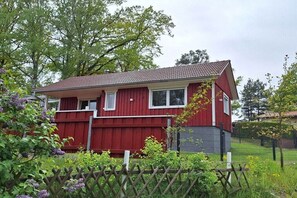 A red two-story house with a tiled roof, surrounded by greenery and a wooden fence. - Bungalow with 1 bedroom (Schwielochsee \/OT Jessern)
