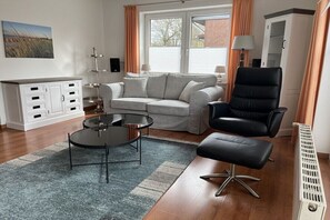 Ein Wohnzimmer mit einer Couch, einem schwarzen Sessel, einem Couchtisch, einem Beistelltisch, einer Lampe, einem Heizkörper und einem Schrank.