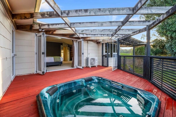 A hot tub on a wooden deck with a covered patio area and a bedroom visible through an open door. - Pokeno Country Retreat | Spa, BBQ and Rural Views (Pōkeno)