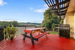 A red wooden deck with a picnic table and grill, overlooking a scenic landscape. - Pokeno Country Retreat | Spa, BBQ and Rural Views (Pōkeno)