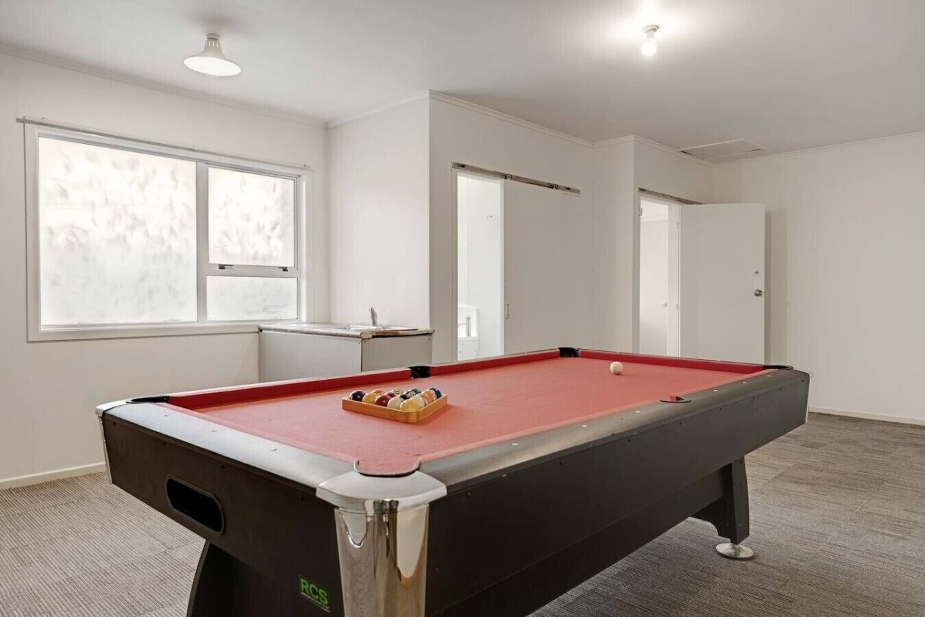 A pool table with balls, a window, and a door in a room with white walls and carpeted floor.