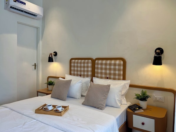 A neatly made bed with a tray of coffee cups and pastries, bedside lamps, a small plant, and a bedside table with a book and a remote control.