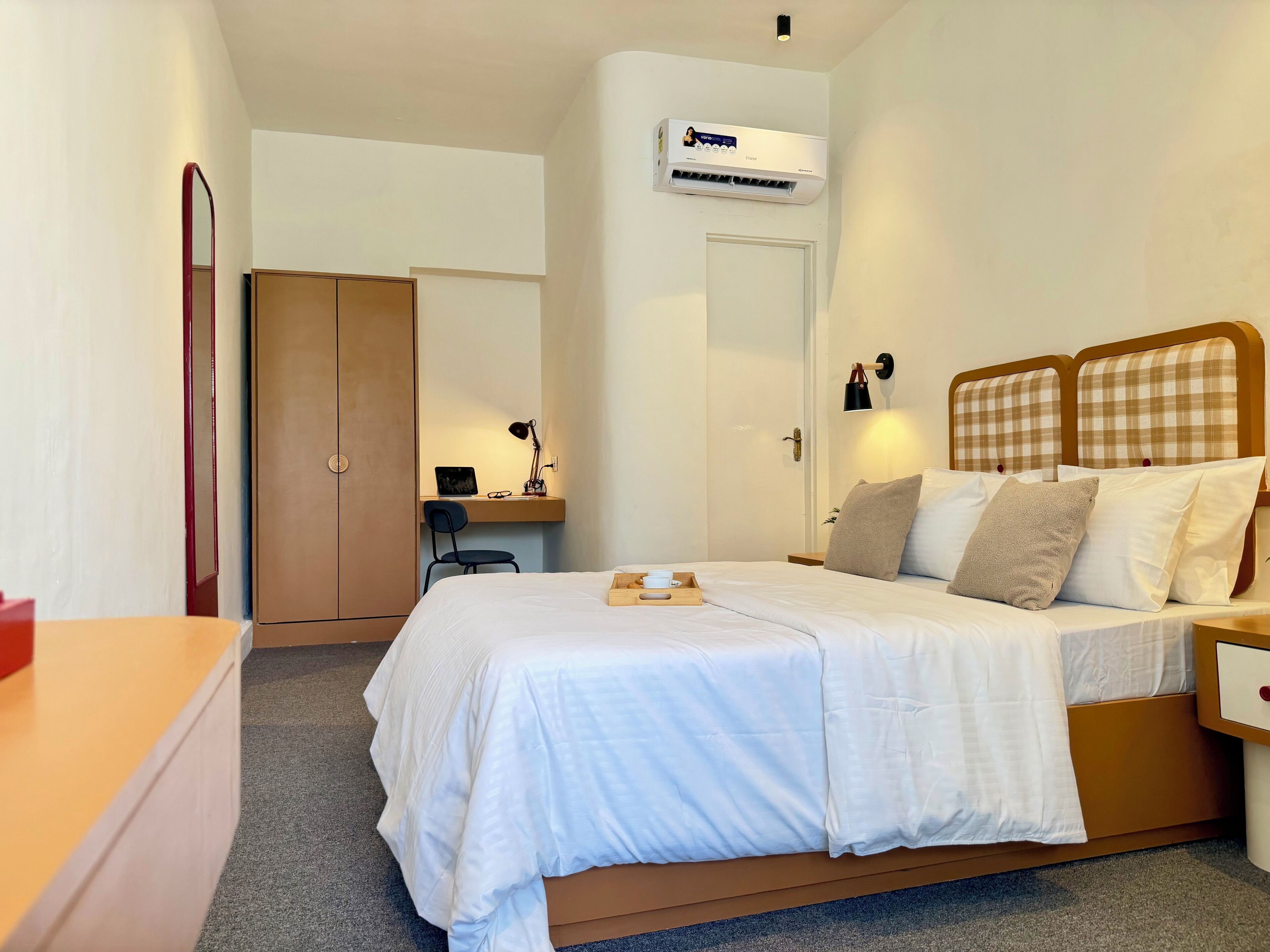 A bedroom with a bed, bedside table, desk, chair, wardrobe, and an air conditioning unit.