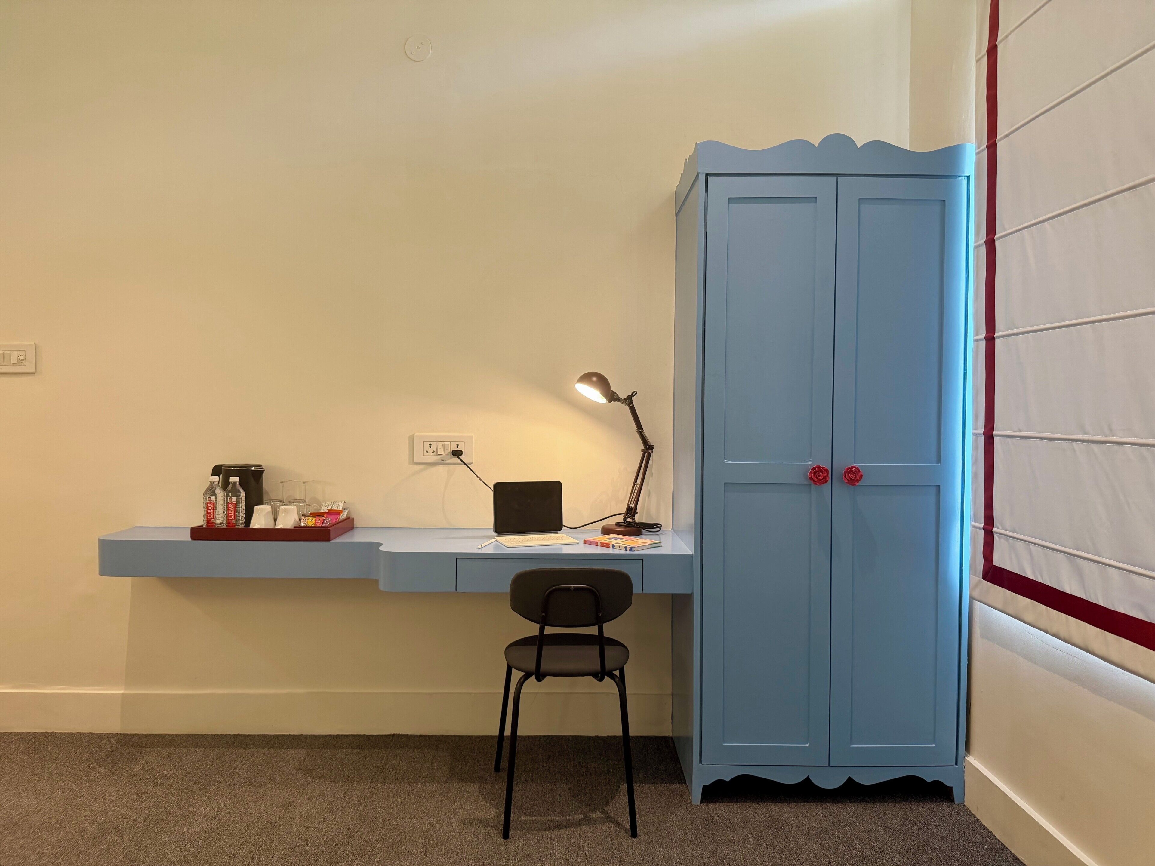 A room with a desk, chair, laptop, lamp, and a blue wardrobe with red handles.