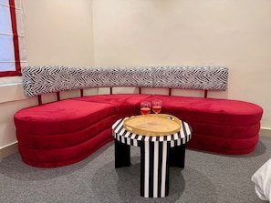 A red upholstered bench with a zebra-striped pillow, a small round table with a striped base, and two glasses of red drink on the table.