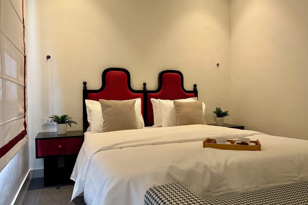 A neatly made bed with white linens and two pillows, a red headboard, a bedside table with a plant, and a tray with a cup and cookies.