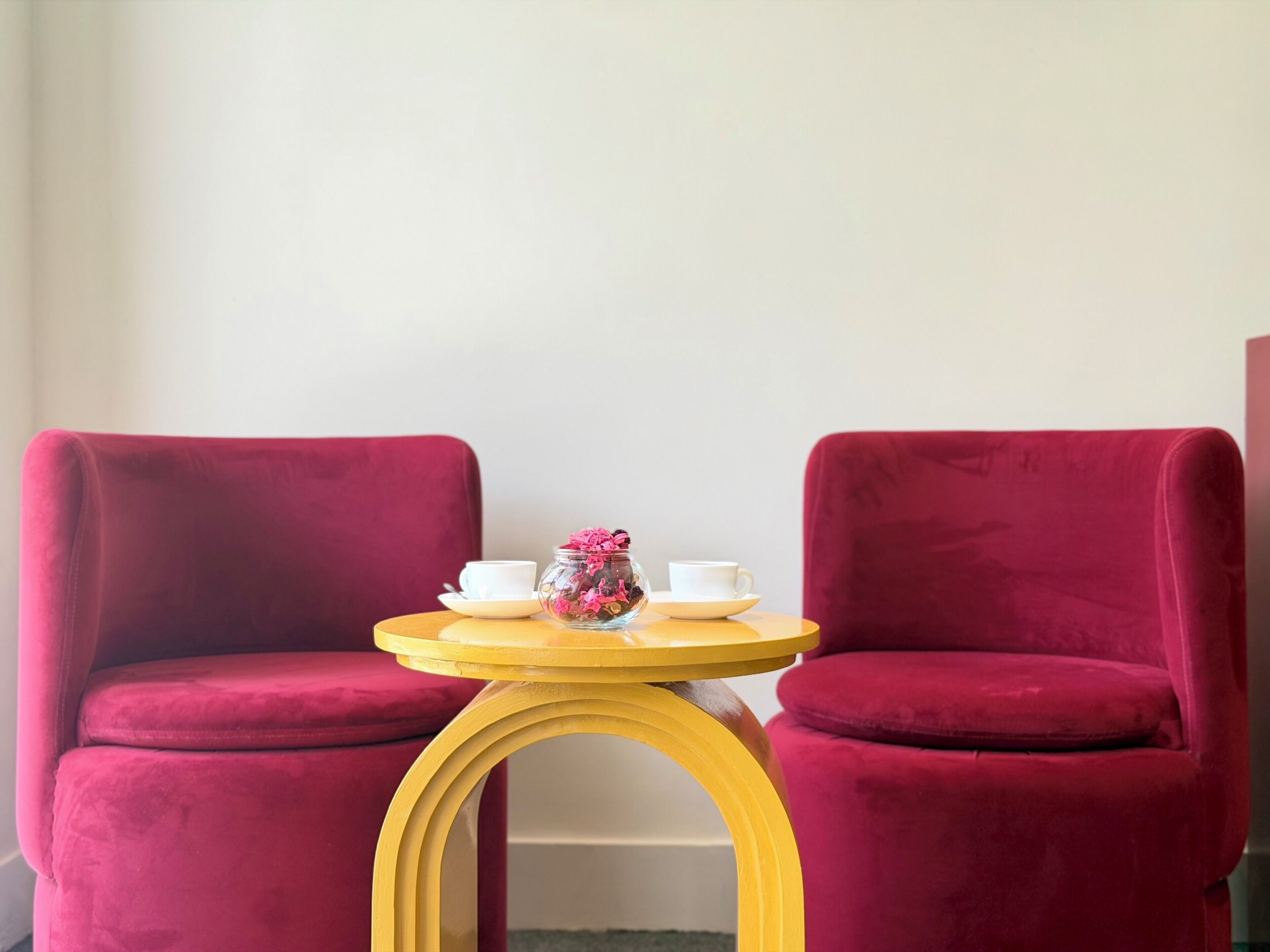 A room with two red upholstered chairs, a small yellow table with a white cup and saucer, and a glass jar with pink flowers.