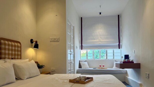A hotel room with a large bed, a bedside table with a lamp, a window with a view of greenery, and a small seating area with a tray of items. - NauKothi Jaipur (Jaipur)