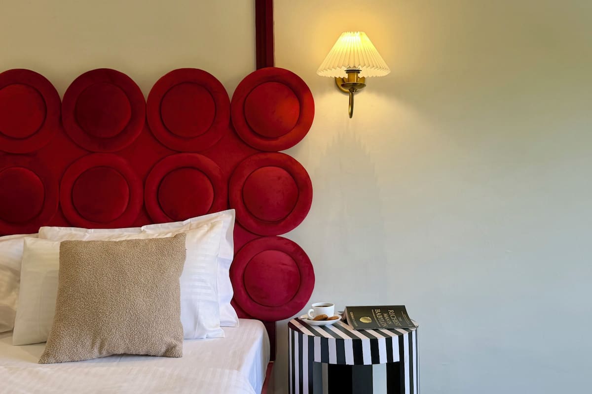 A neatly made bed with pillows, a red headboard, a bedside lamp, and a small table with a book and a cup.