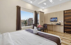 Et hotelværelse med seng, skrivebord, fjernsyn og et vindue med gardiner.