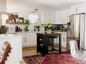 A modern kitchen with white cabinets, a stainless steel refrigerator, a round mirror, and a central island with stools. - Caviar on Pine in Downtown McKinney • Sleeps 9 • Luxe Stay • Elevated Coffee Bar (McKinney)