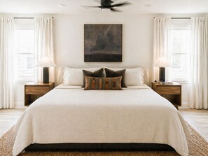 Ein großes Bett mit weißer Bettdecke, zwei Nachttische mit Lampen, ein Deckenventilator und ein Gemälde an der Wand.