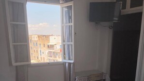 A room with two beds, a table with a patterned tablecloth, a TV mounted on the wall, and a window with curtains. - Ritza Hotel (Tunis)
