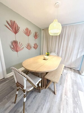 Un coin repas avec une table ronde, des chaises en osier, un luminaire suspendu et un mur orné de motifs de feuilles.