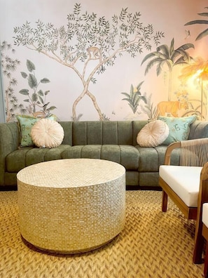Ein grünes Sofa mit gemusterten Kissen, ein gewebter runder Tisch und ein Wandbild mit Baum und tropischer Szene.