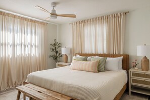 Een slaapkamer met een bed, nachtkastjes, een bankje, een plafondventilator en dunne gordijnen.