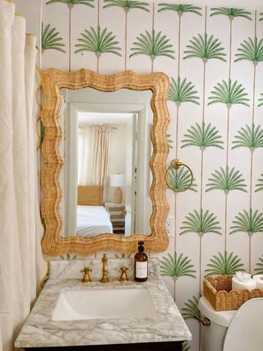 Une salle de bain avec un lavabo en marbre, un miroir encadré de rotin et des toilettes accompagnées d’un panier en rotin.