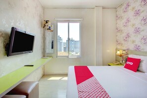 Quarto de hotel com cama, televisão, escrivaninha, janela com vista e mesa de cabeceira com abajur.