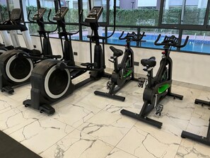 Ett gym med motionscyklar och crosstrainers, stora fönster och utsikt över poolen.