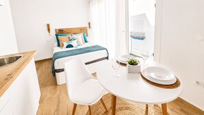 A modern hotel room with a bed, a dining table with chairs, a small kitchen area, and a window with a balcony. - CREA Conil Apartments III (Conil de la Frontera)