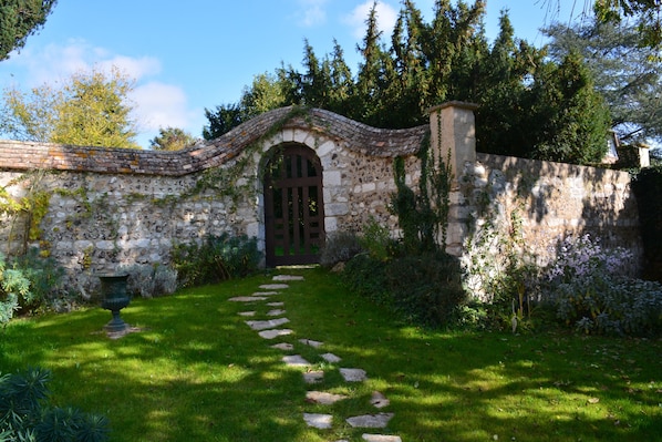 Un arc en pierre mĂšne Ă un jardin oĂč serpente un sentier dallĂ©.