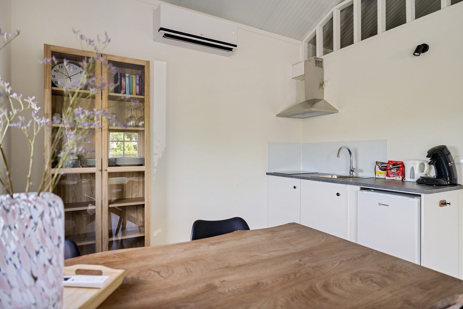 A compact kitchen with a wooden table, a built-in bookshelf, a sink, and a coffee machine.