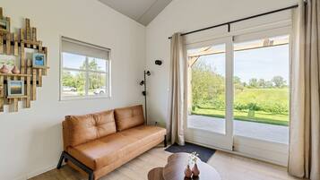 Ein Zimmer mit einer braunen Ledersofa, einem kleinen runden Tisch mit Vasen, einer Stehlampe und Blick auf eine grüne Landschaft durch große Schiebeglasfenster.