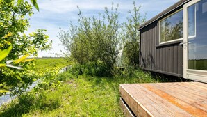 A wooden deck adjacent to a building with a large window, surrounded by greenery and a small water channel. - Camping de Wartenster - Pipowagen (Wartena)