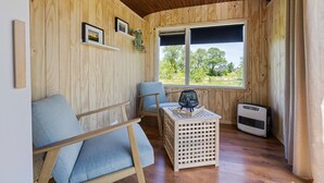 A wooden-paneled room with a window, a chair, a small table, and a heater. - Camping de Wartenster - Pipowagen (Wartena)
