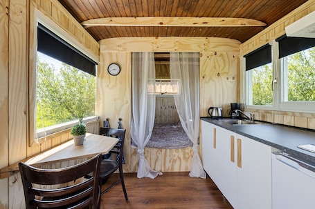 A compact room with a bed, a dining table, chairs, a sink, and a clock on the wall.. Camping de Wartenster - Pipowagen