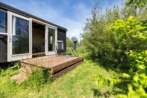 A modern mobile home with a deck, large windows, and a view of the surrounding greenery.