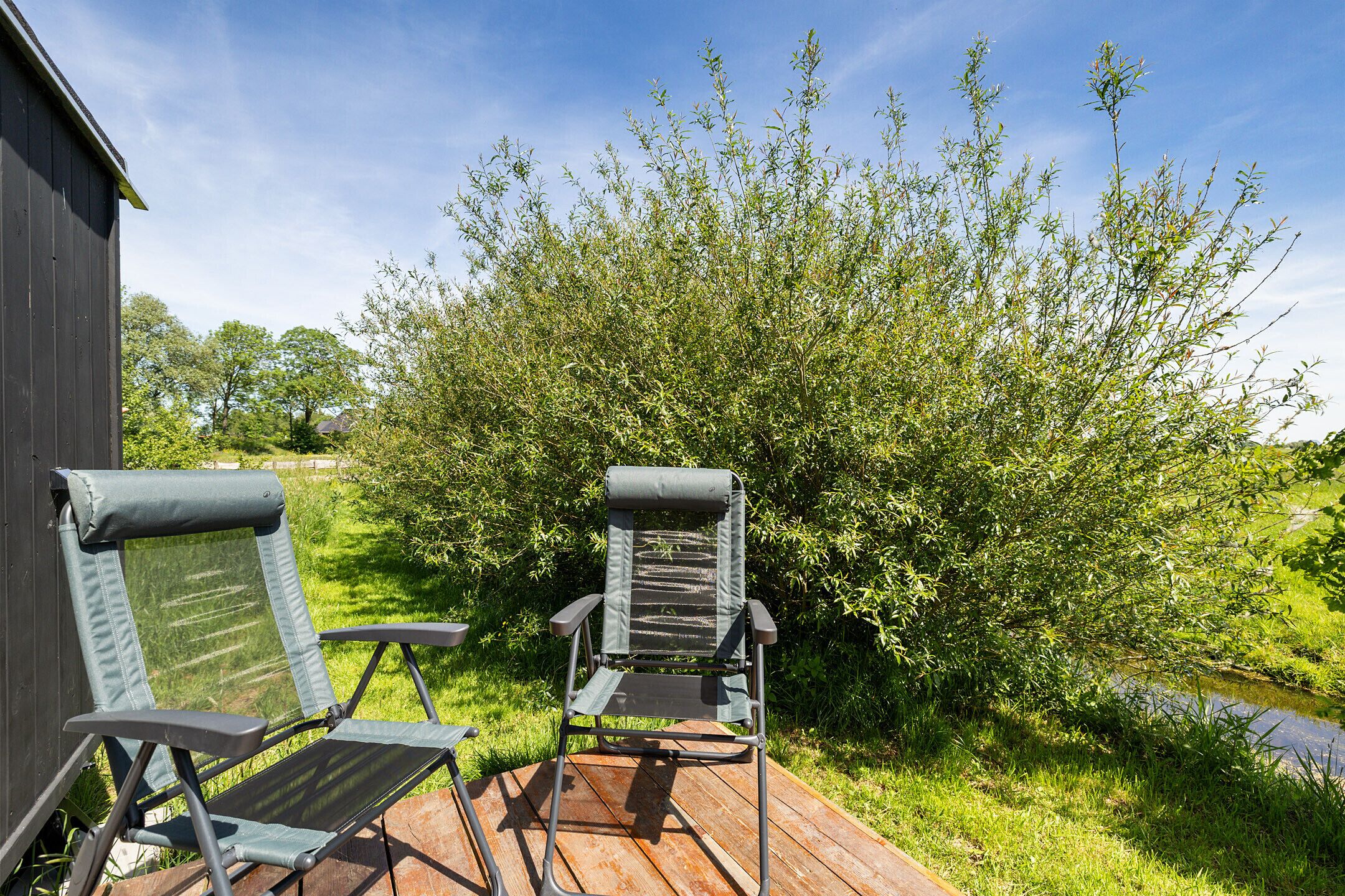 Two outdoor chairs on a wooden deck with a view of a garden and a building in the background.