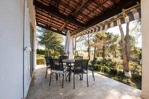 Un patio al aire libre con mesa y sillas, una pérgola arriba y vistas a un jardín con árboles y arbustos.