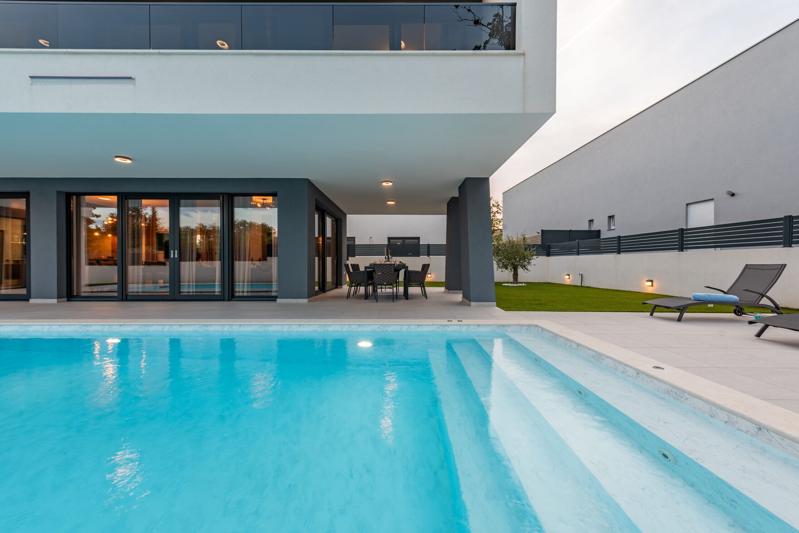 Un espace piscine extérieur moderne, avec une piscine à l’eau d’un bleu limpide, un coin repas aménagé avec du mobilier d’extérieur, et un grand bâtiment doté d’un toit plat et de grandes fenêtres.