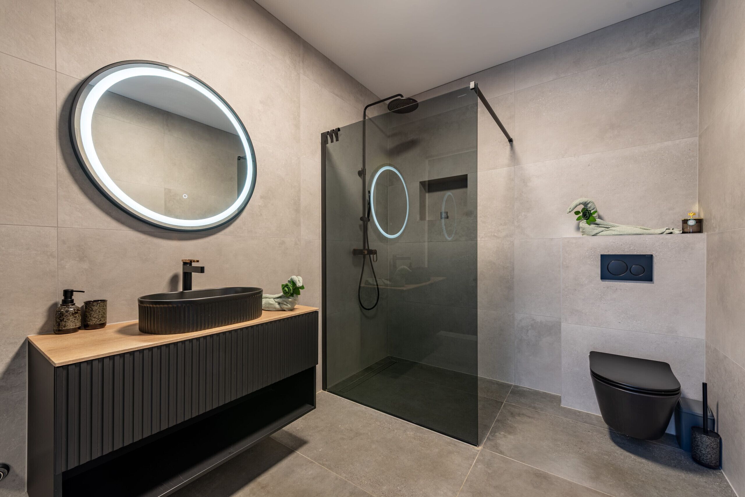 A modern bathroom with a round mirror, a glass-enclosed shower, a wooden vanity, and a toilet.