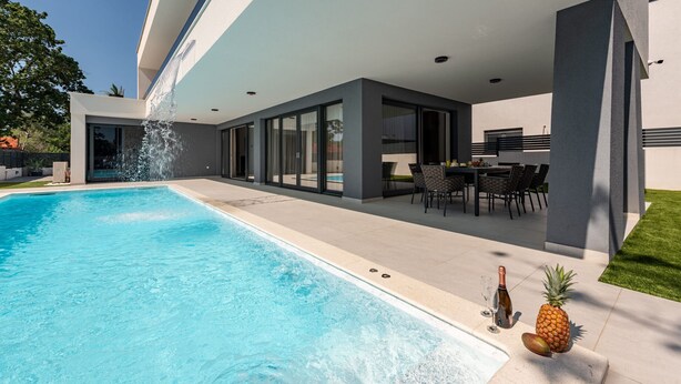 Un área moderna de piscina al aire libre con una piscina azul cristalina, un elemento acuático y un espacio de comedor con mobiliario exterior.