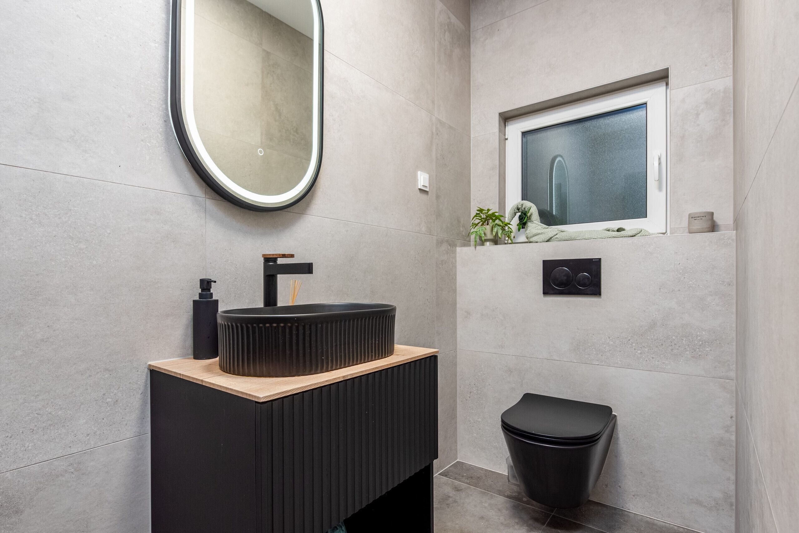 A modern bathroom with a round mirror, a black sink, a window, and a black trash bin.