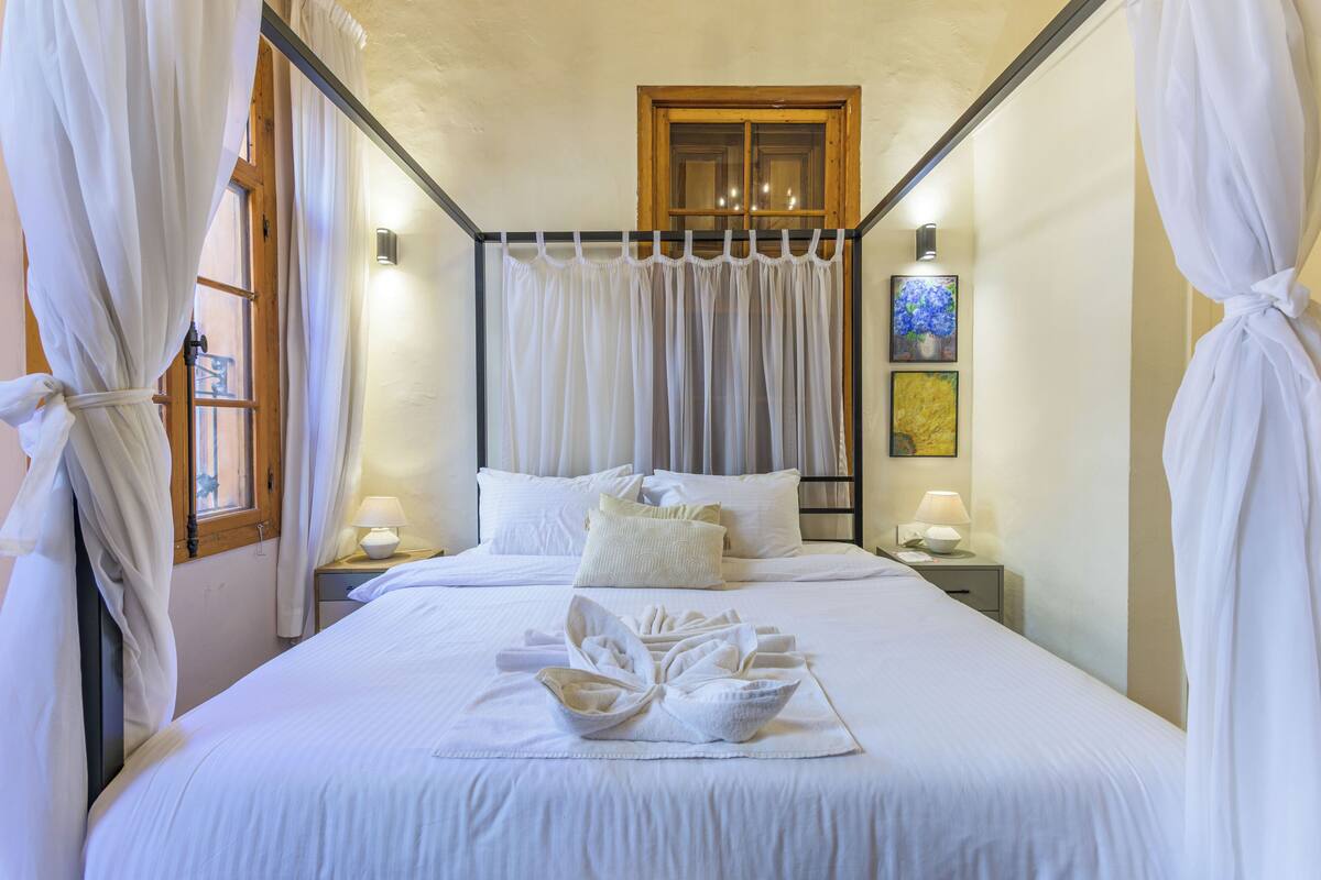 Una cama bien tendida con sábanas blancas y un arreglo de toallas, una ventana con marco de madera y dos lámparas de noche.