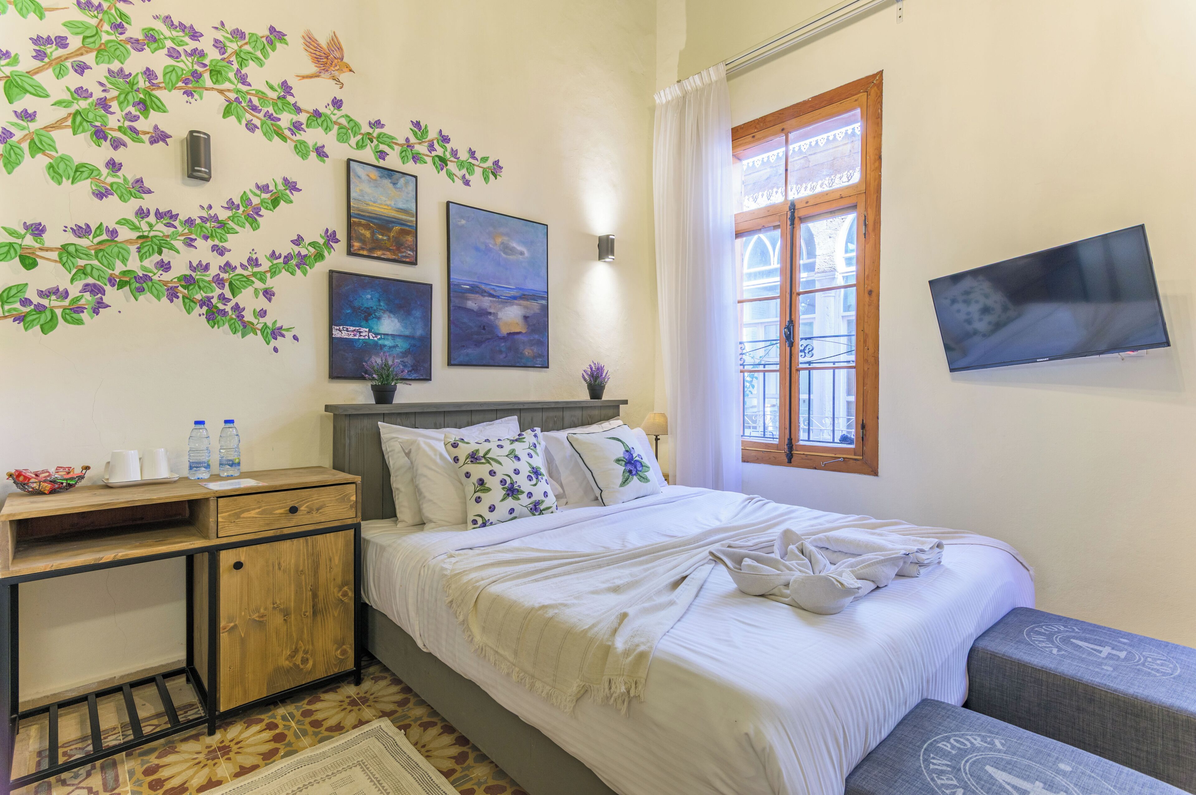 A bedroom with a bed, a nightstand, a desk, a window, and wall art.