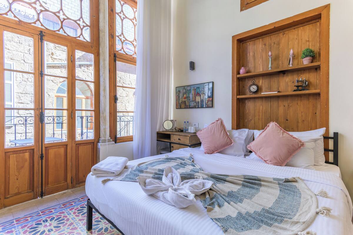 Habitación de hotel con cama, armario de madera, escritorio con silla, ventana con cortinas y un cuadro de un paisaje urbano en la pared.