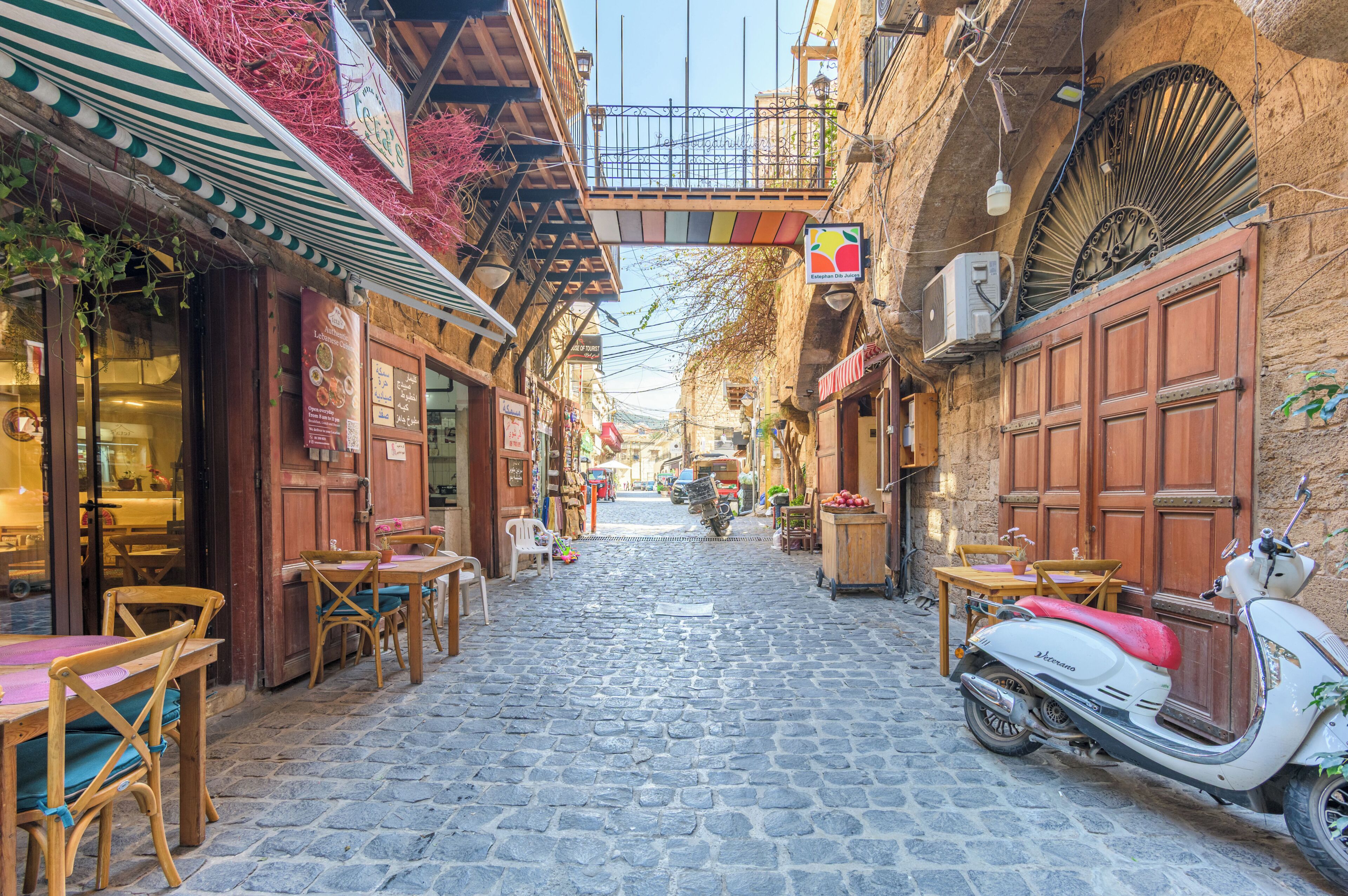 Une rue pavée avec un scooter garé, des tables et des chaises en bois, et un bâtiment avec une enseigne.