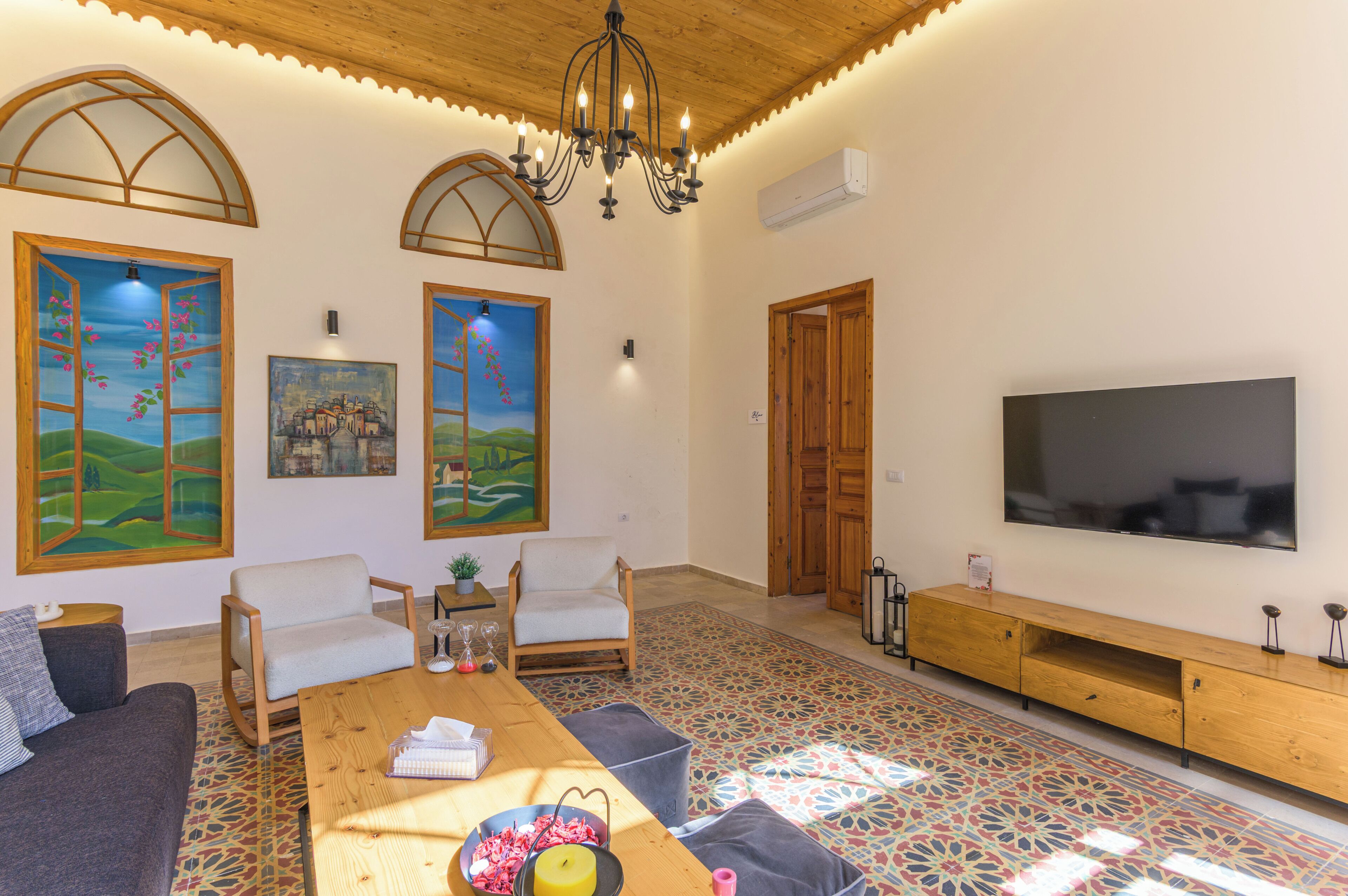A living room with a wooden ceiling, a large TV, two white chairs, a wooden coffee table, and colorful floor tiles.