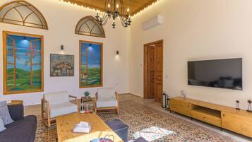 A living room with a wooden ceiling, a large TV, two white chairs, a wooden coffee table, and colorful floor tiles.