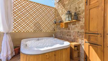 A wooden bathtub in a stone-walled room with a lattice screen, a wooden door, and a shelf with decorative items.