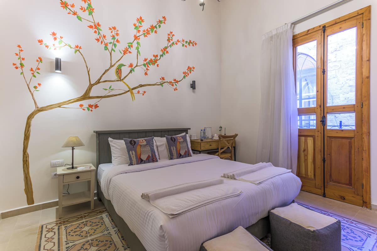 Un dormitorio con cama, mesita de noche, lámpara, mesita de madera, banco, estante en la pared y ventana con cortinas.