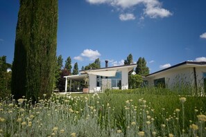 A modern house with a covered patio, surrounded by tall trees and a field of flowers. - Nestled among the gentle hills of San Marcello, where the eye wanders across lush greenery and the silhouette of the mountains, stands a private villa designed for those who wish to slow down, breathe deeply, and enjoy the luxury of time.Offering 500 sqm (San Marcello)