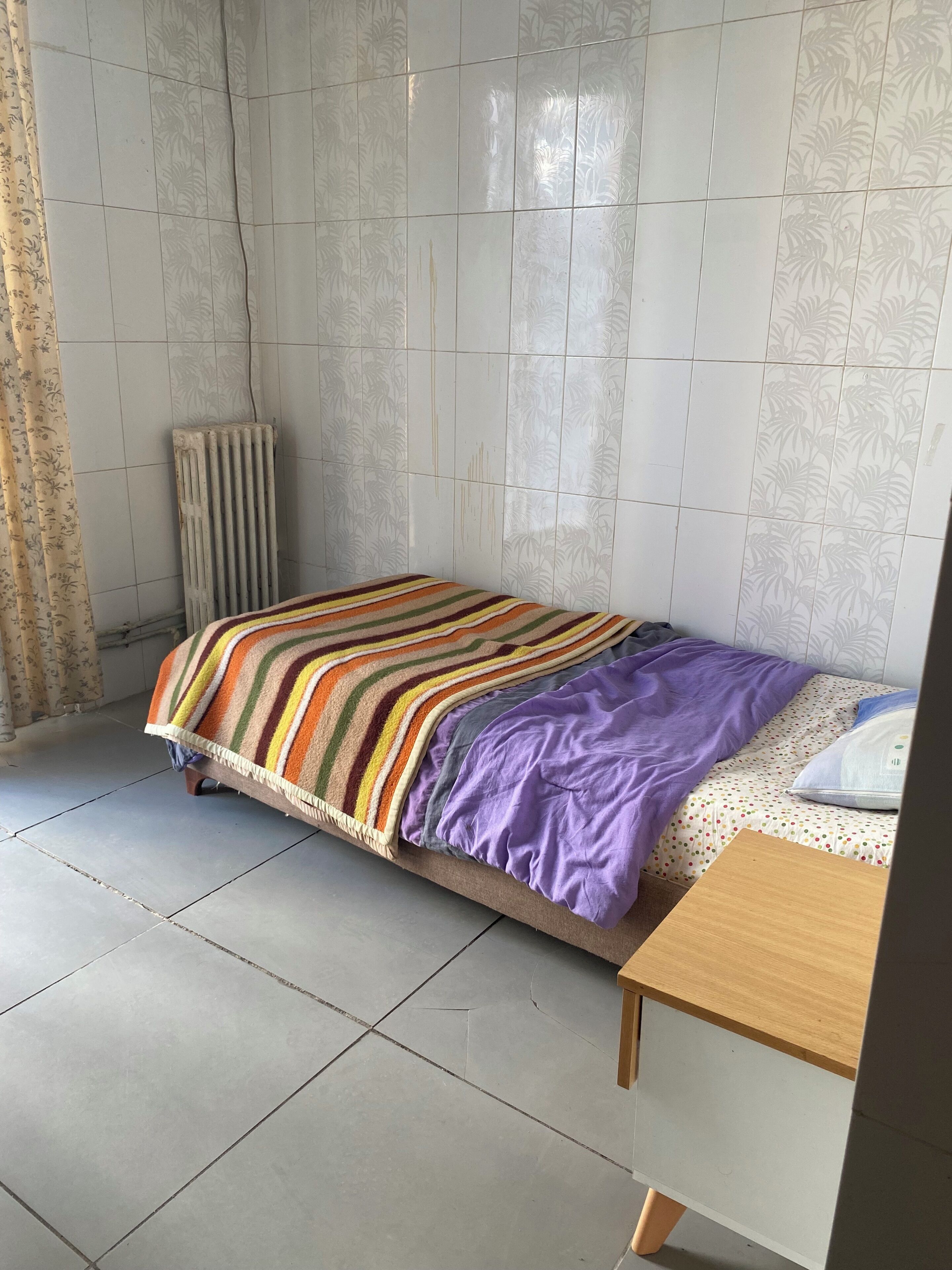 Un lit simple avec une couverture à rayures, une table de chevet en bois et un radiateur adossé à un mur carrelé.