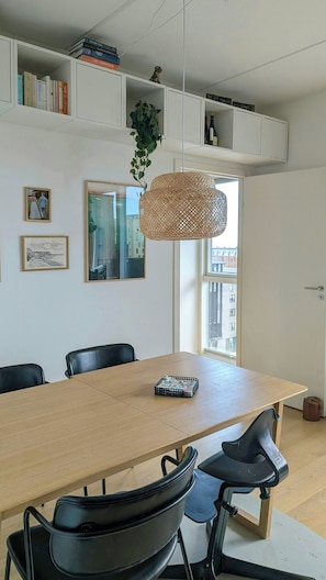 Ein moderner Essbereich mit einem langen Tisch, schwarzen Stühlen, einer gewebten Pendelleuchte und einem Bücherregal darüber.